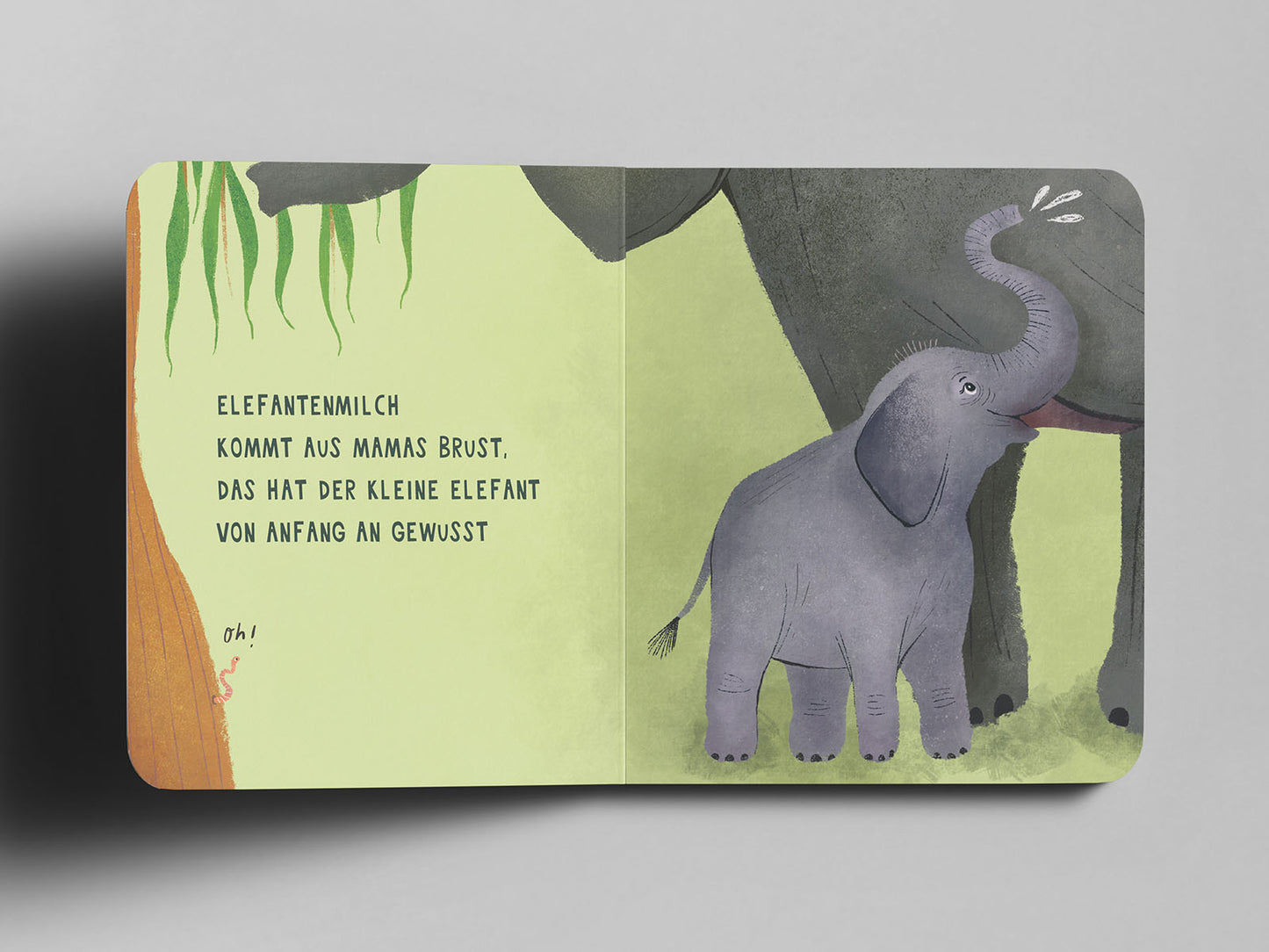 Aufgeschlagenes Pappbuch mit einer Illustration eines kleinen, lilagrauen Elefanten, der an der Brust seiner großen Elefantenmama trinkt. Links im Bild hängen grüne Blätter herab, am unteren Rand schlängelt sich ein kleiner Wurm mit dem handschriftlichen Text ‚oh!‘ daneben. Auf der linken Seite steht in schwarzer Schrift: ‚Elefantenmilch kommt aus Mamas Brust, das hat der kleine Elefant von Anfang an gewusst‘.