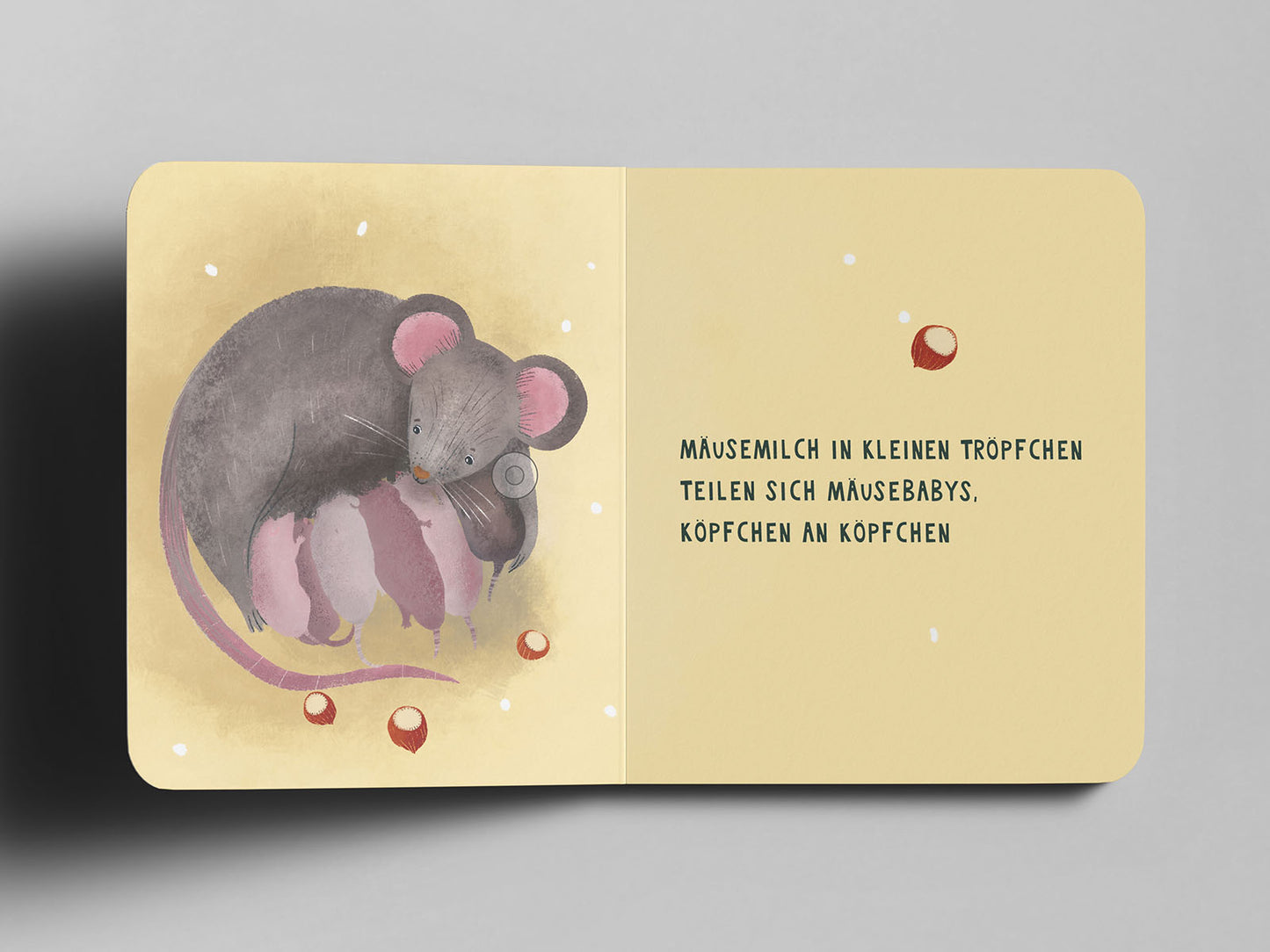 Aufgeschlagenes Pappbuch mit einer Illustration einer grauen Maus, die liebevoll ihre rosa Mäusebabys säugt. Die Babys liegen eng aneinander, Köpchen an Köpchen. Kleine, Haselnüsse sind um sie herum verteilt. Der Hintergrund ist warm beige mit weißen Sprenkeln. Auf der rechten Seite steht in schwarzer Schrift: ‚Mäusemilch in kleinen Tröpfchen teilen sich Mäusebabys, Köpfchen an Köpfchen‘.