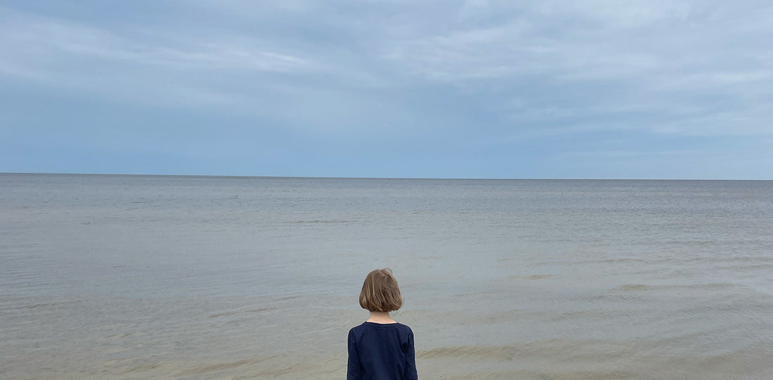 Weiter Horizont am Meer. Ein Kind mit mittellangem Haar steht mit dem Rücken zur Kamera und blickt in die Ferne.