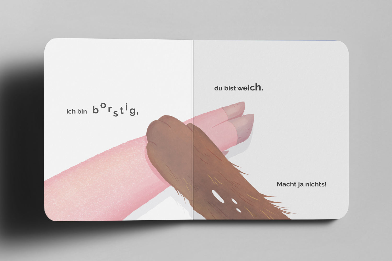 Kinderbuch-Doppelseite mit einer Nahaufnahme eines rosa Schweinebeins und einer braunen Hundepfote, die sich sanft berühren. Der Hintergrund ist weiß. Links steht der Text „Ich bin borstig,“ mit hervorgehobenen Buchstaben. Rechts folgt „du bist weich.“ in fetter Schrift und darunter kleiner „Macht ja nichts!“.