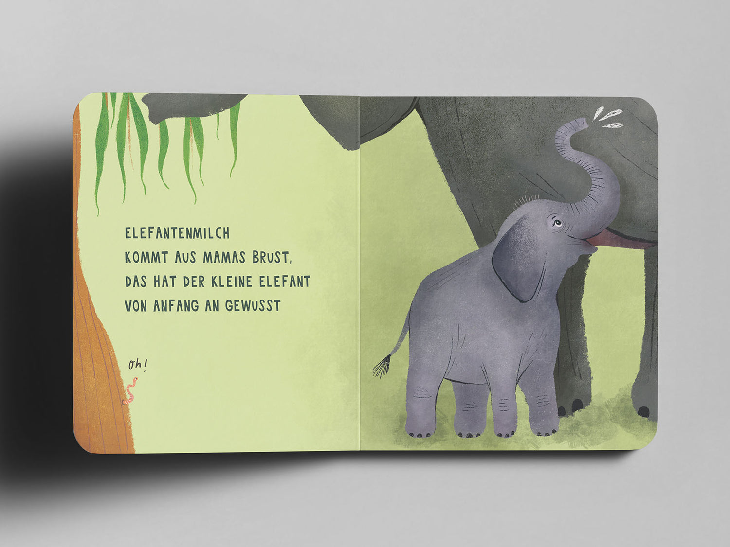 Aufgeschlagenes Pappbuch mit einer Illustration eines kleinen, lilagrauen Elefanten, der an der Brust seiner großen Elefantenmama trinkt. Links im Bild hängen grüne Blätter herab, am unteren Rand schlängelt sich ein kleiner Wurm mit dem handschriftlichen Text ‚oh!‘ daneben. Auf der linken Seite steht in schwarzer Schrift: ‚Elefantenmilch kommt aus Mamas Brust, das hat der kleine Elefant von Anfang an gewusst‘.